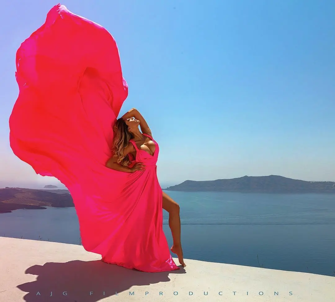 Bright pink flowing-dress fashion image against coastal architecture.
